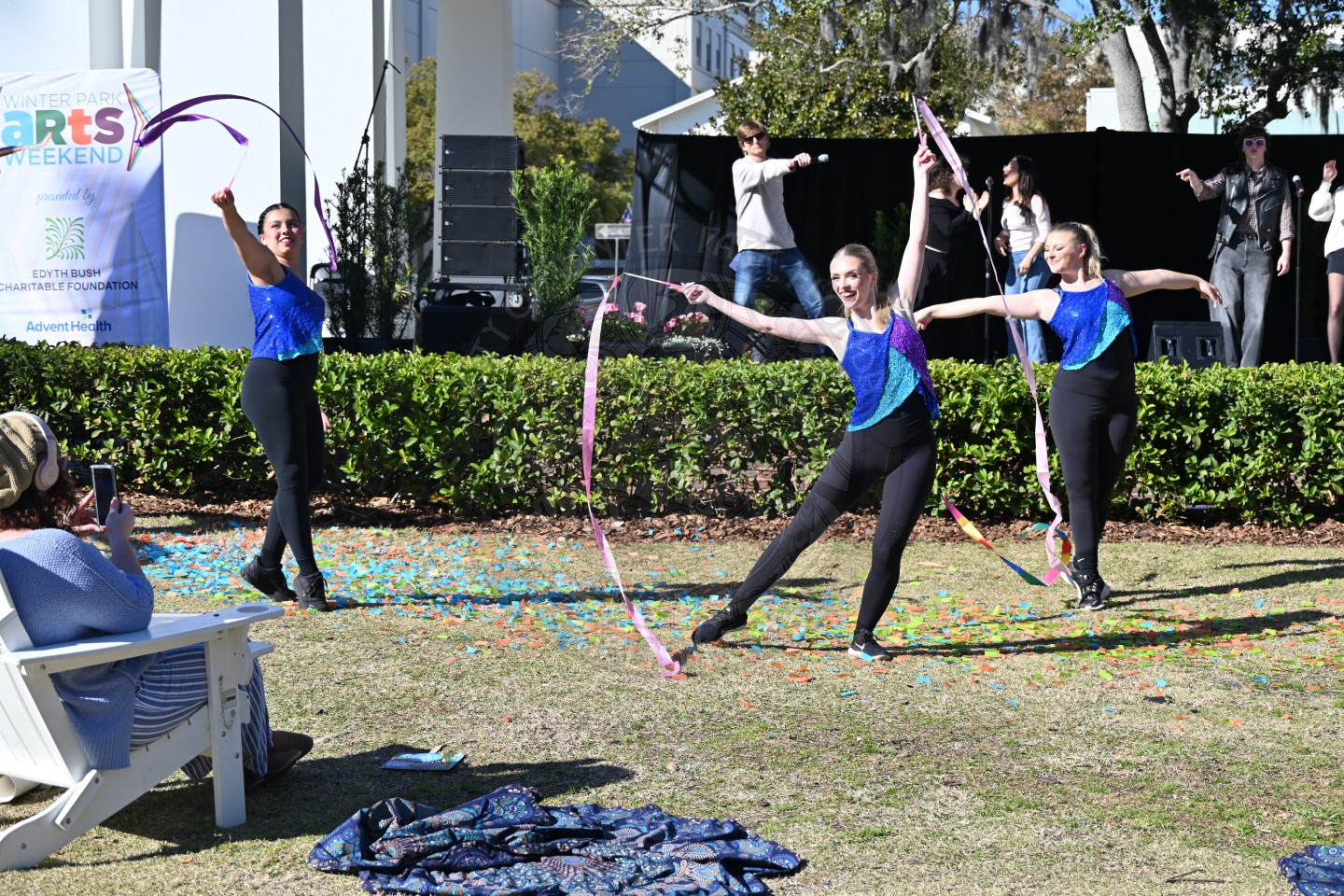 Three women in matching blue and black outfits perform a ribbon dance outside during a Winter Park Arts event, with an audience member seated in front and other performers and a stage in the background.