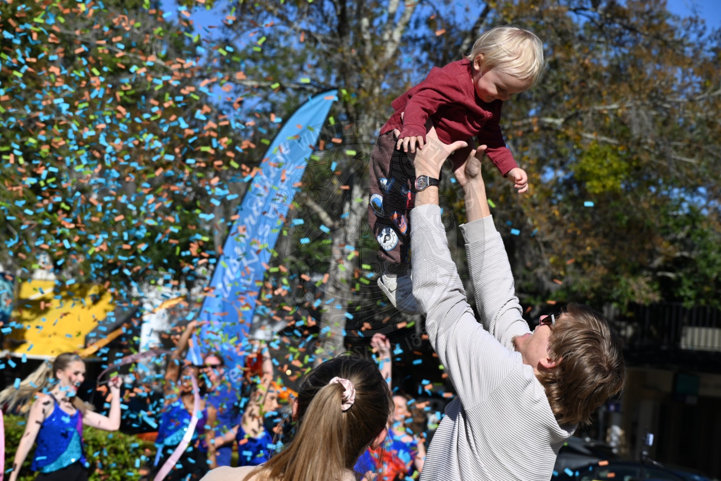 A man lifts a smiling toddler into the air as confetti falls around them at an outdoor celebration. A woman with a ponytail stands nearby, and people in colorful outfits cheer in the background.