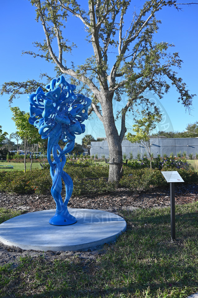 Abstract sculpture mounted on a round concrete pedestal in a landscaped park setting.