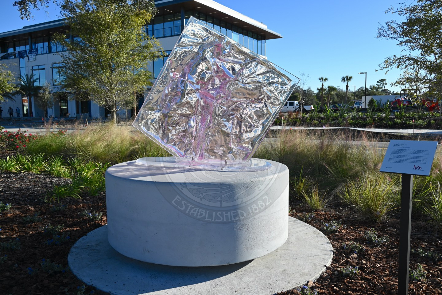 Abstract glass sculpture mounted on a round concrete pedestal in a landscaped park setting with a building in the background.