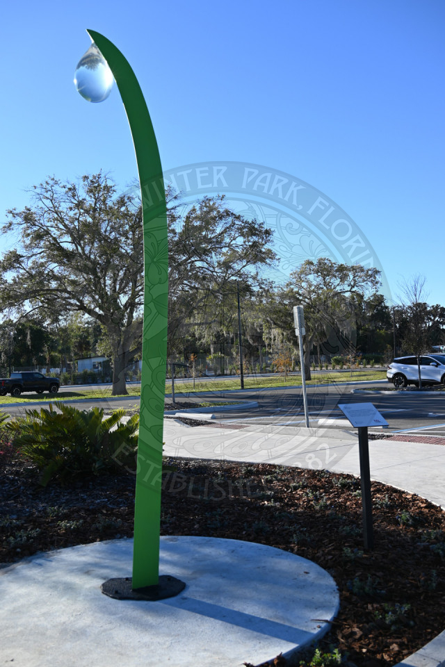 Abstract sculpture mounted on a round concrete pedestal in a landscaped park setting with a parking lot in the background.
