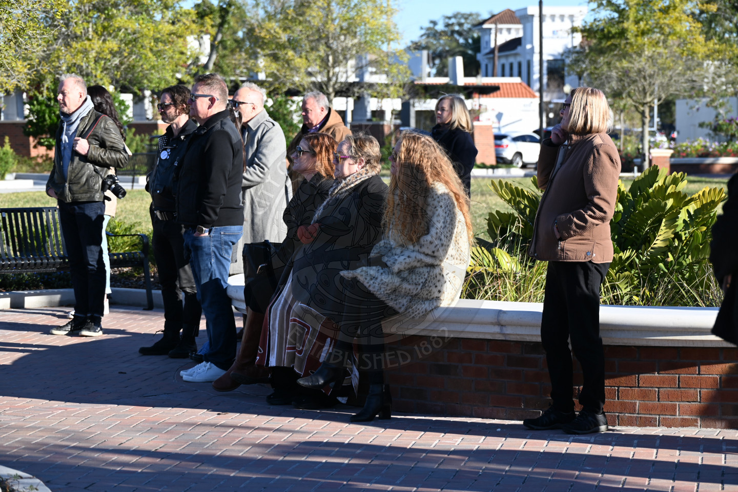 Group of people gathered in a sunny park listening to a speaker during an outdoor event near benches and landscaping.