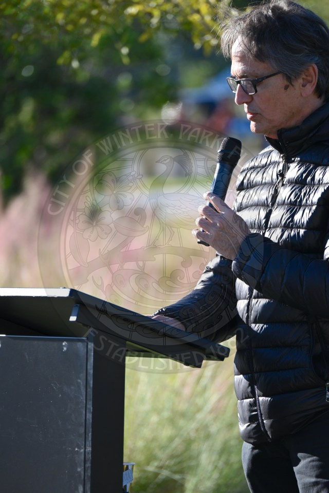 Man speaking at a podium outdoors while reading from notes and holding a microphone.
