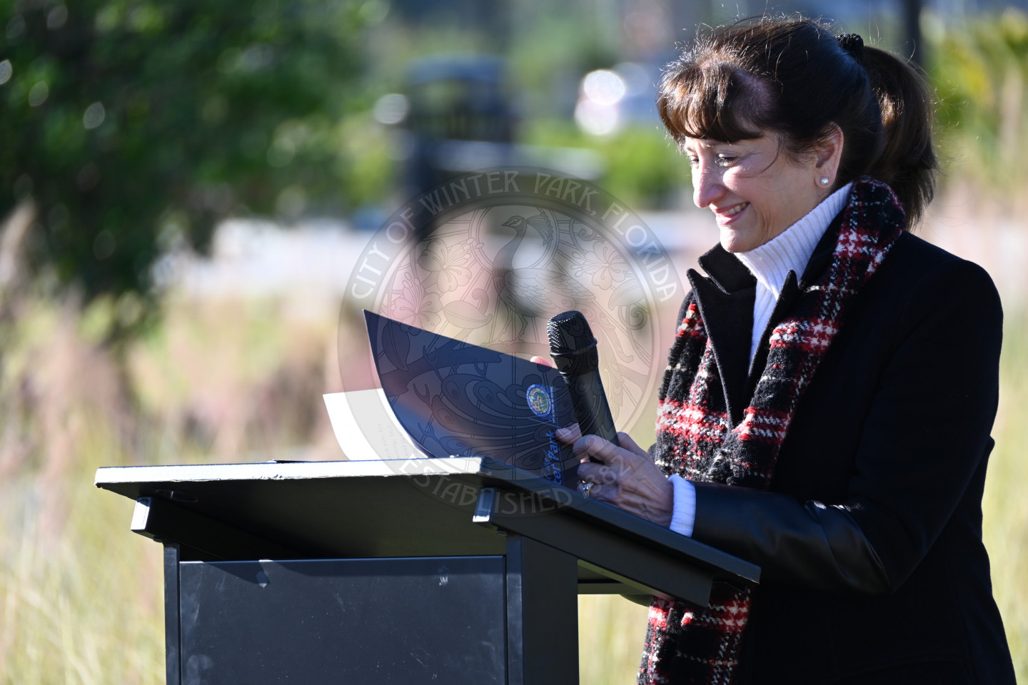 Woman speaking at a podium outdoors while reading from notes and holding a microphone.