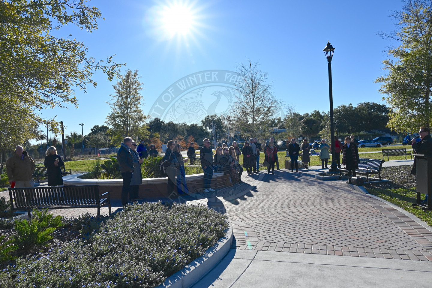 Group of people gathered in a sunny park listening to a speaker during an outdoor event near benches and landscaping.