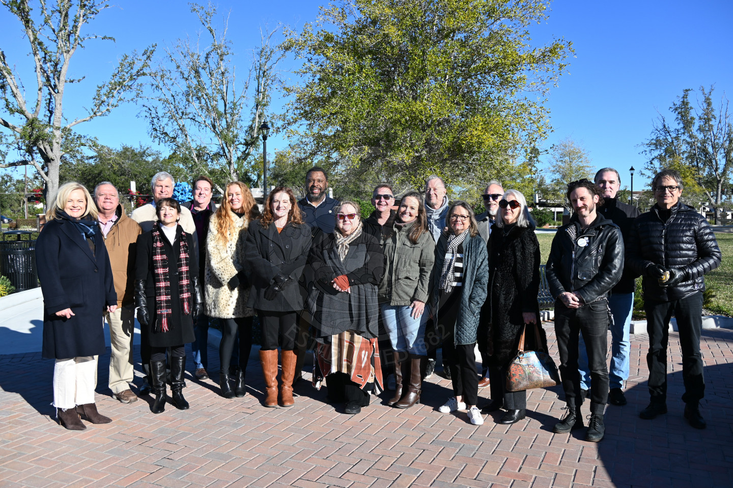 a big group of people standing next to one another posing for a picture in a sunny park with a live oak tree behind them.