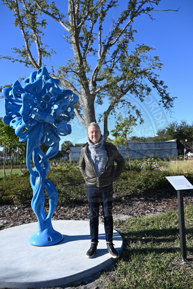 An artist standing next to his abstract sculpture with a tree and building in the background.