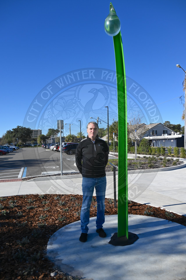 An artist standing next to his abstract sculpture with a parking lot and buildings in the background.