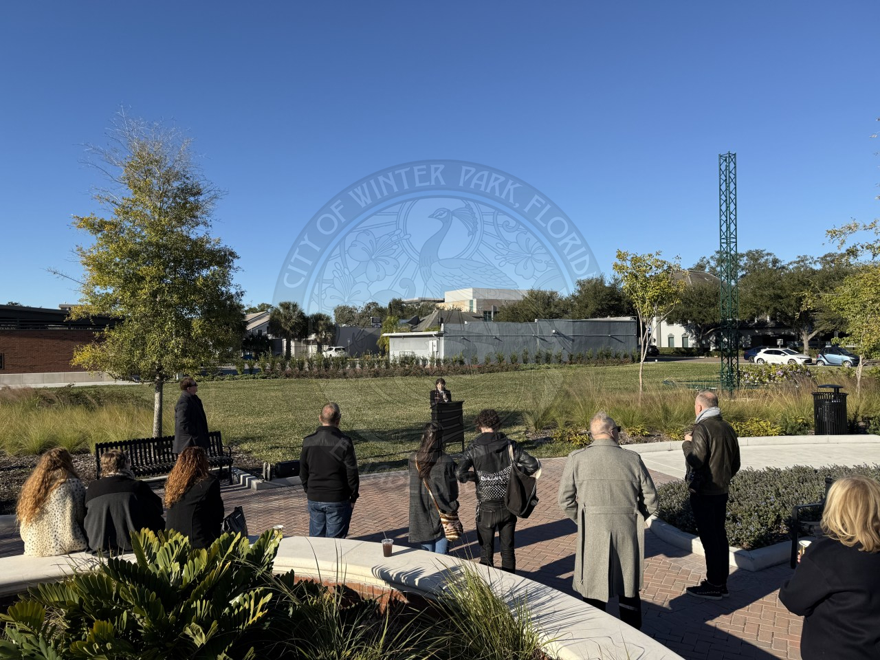 Group of people gathered in a sunny park listening to a speaker during an outdoor event near benches and landscaping.