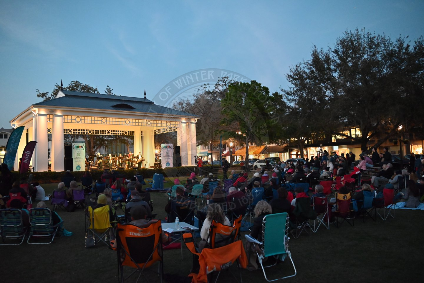 a bunch of people sitting on the grass and in lawn chairs in front of a stage listening to the orchestra perform.