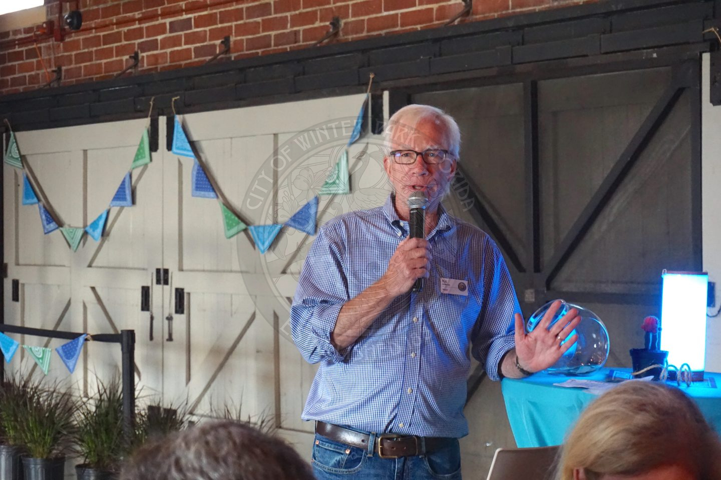 a man leaning his left forearm on a hightop table and speaking into a wireless microphone with his right hand