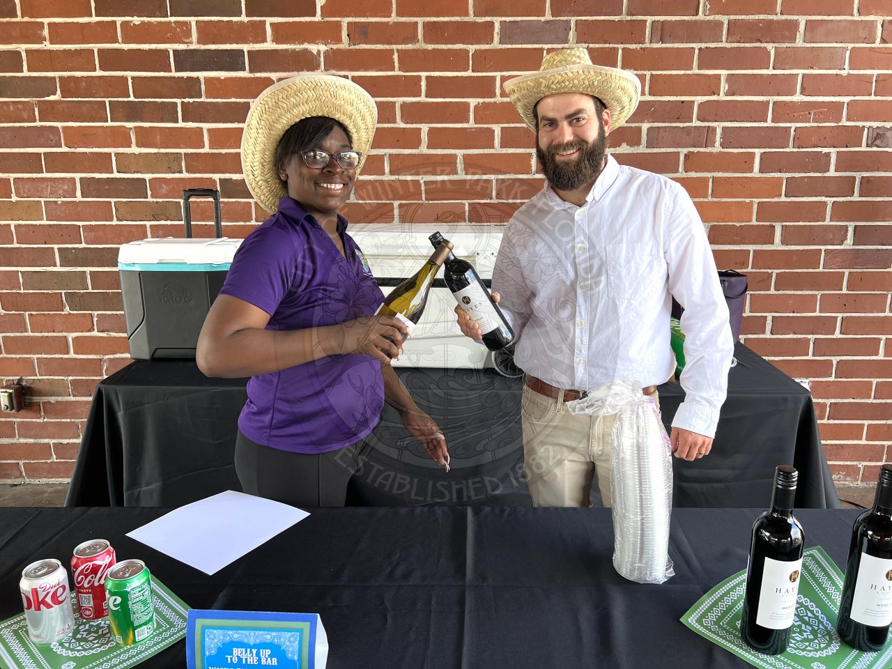 a man and woman wearinc cowboy hats standing behind a table while both of them are holding wine bottles smiling