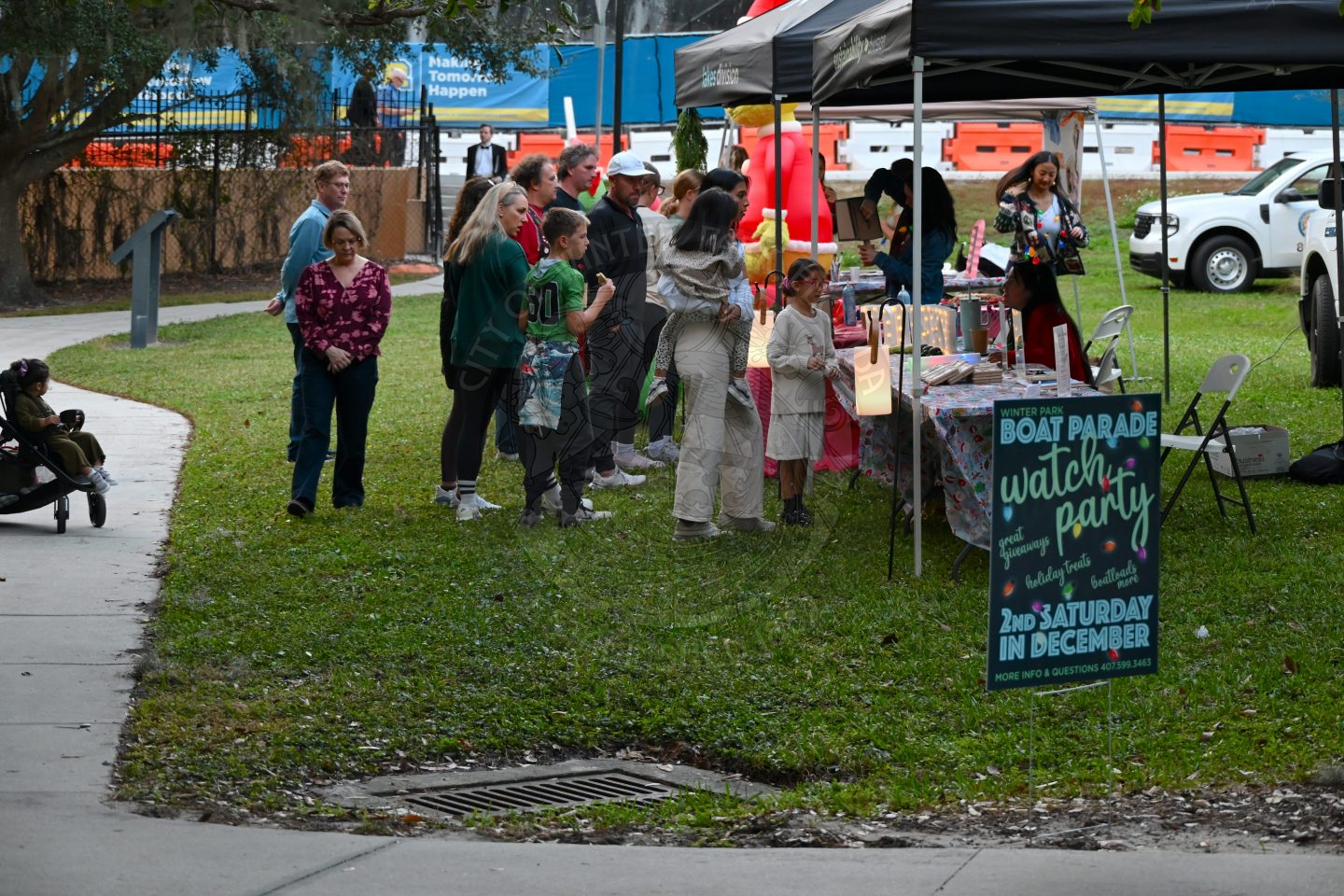 people standing in front of tables with tents covering the tables. There is a sign that says "Winter Park Boat Parade Watch Party 2nd Saturday in December"