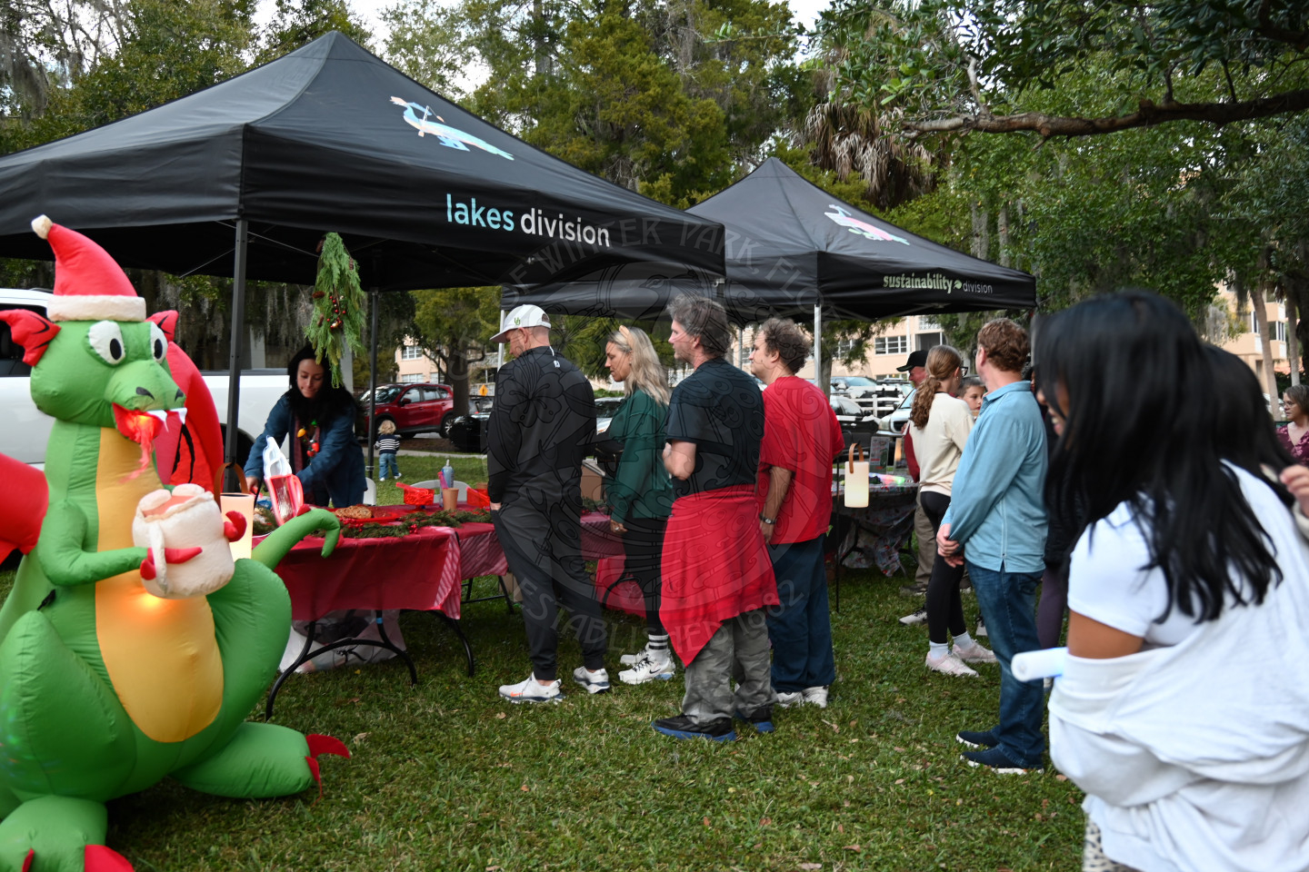 People standing in a line in front of tables with tents covering the tables. To the left of the tents is a blow up dragon wearing a santa hat.