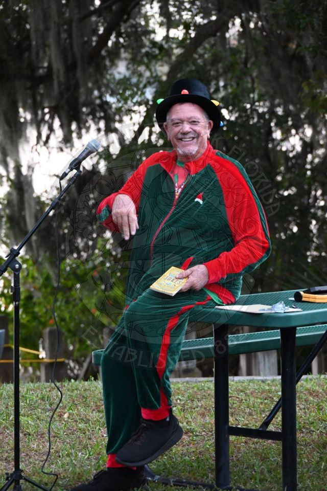 A man sitting on a picnic table wearing a red and green velour sweat suit with a top hat with Christmas lights.