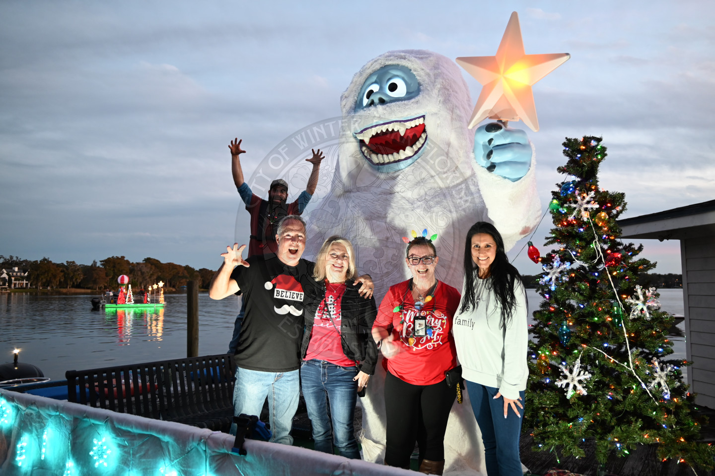 Four adults smiling and posing, standing in front of a large white abominable snowman holding a glowing star. One person behind them raises their arms playfully. A decorated Christmas tree with colorful lights stands to the right.