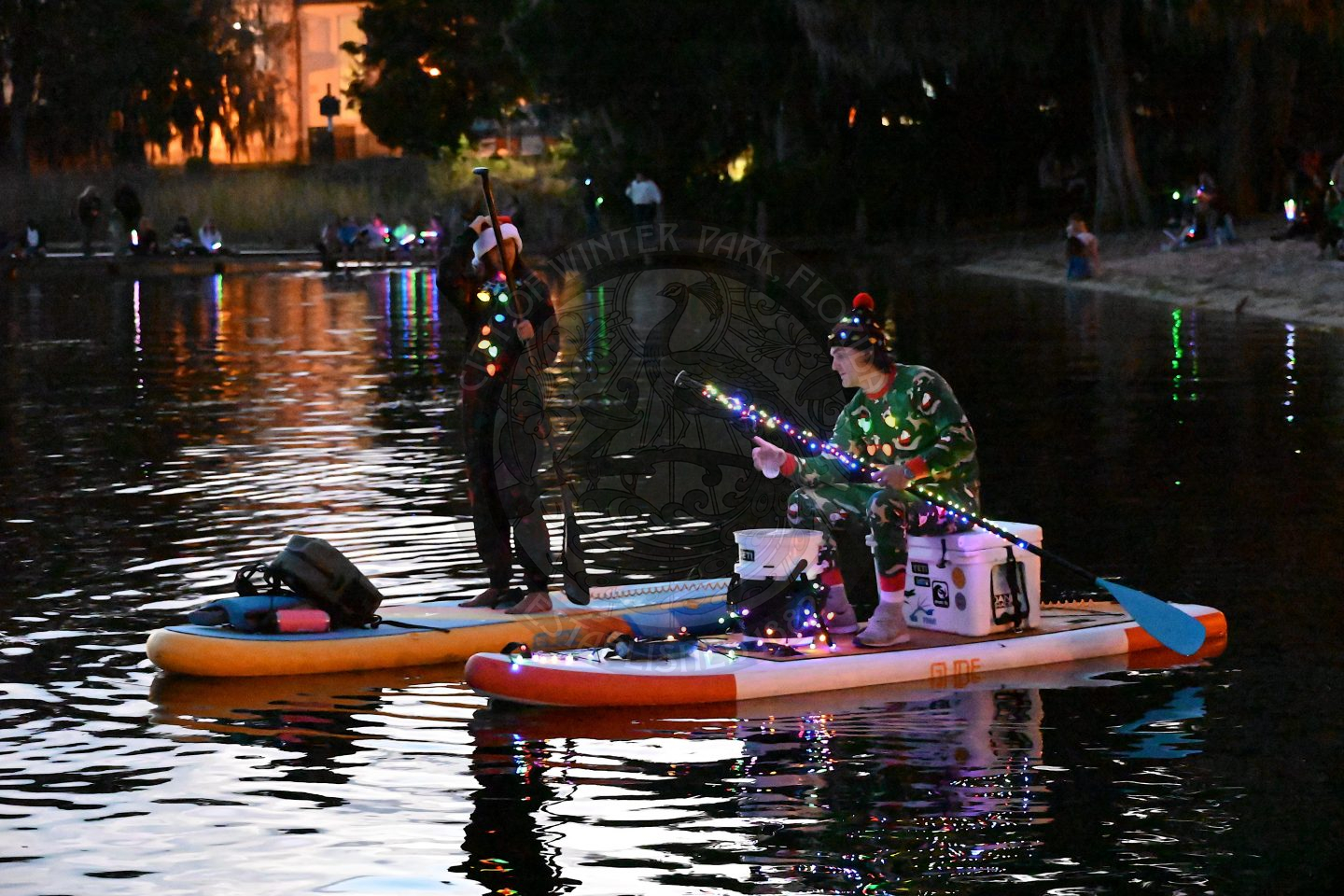 two people with paddleboards on water. One guy is sitting on a cooler that is on the paddleboard and the girl is standing on her paddleboard.