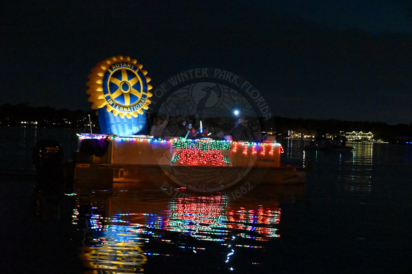 A boat cruising at night, brightly decorated with colorful holiday lights and a blow up that says "Winter Park Rotary"