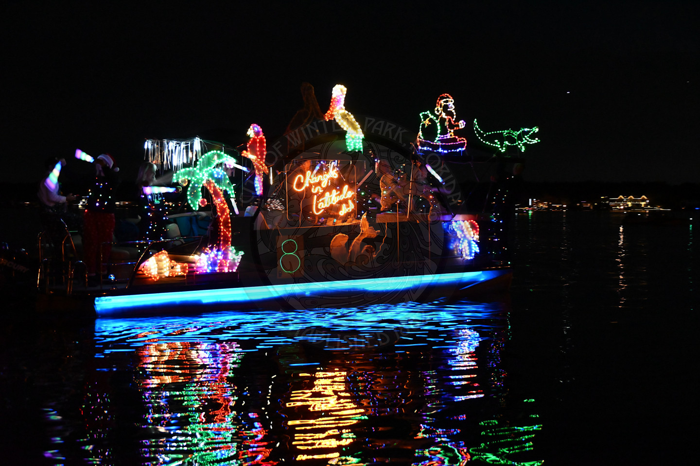 A boat at night, brightly decorated with colorful holiday lights. Lighted figures include palm trees, pelican, parrot, and a gator with a glowing sign.