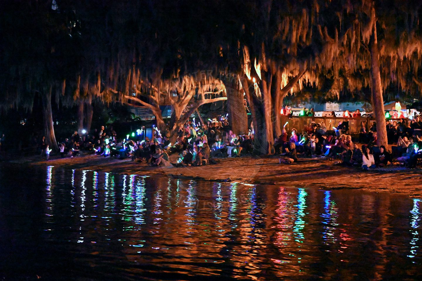a bunch of people sitting lakeside wearing light ups necklaces, hats, bracelets, etc. watching all the boats in the water.