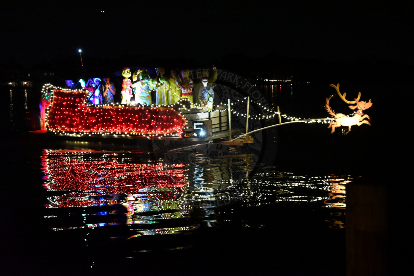 A boat cruising at night, brightly decorated with colorful holiday lights. Has red lights in the shape of a sleigh with light up grinch on the boat. There is Max from the movie grinch on a pole sticking out in front.