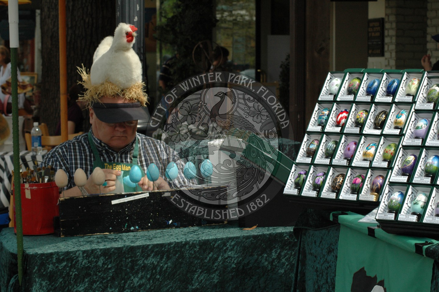 Male vendor wearing a hat with a stuffed chicken on the hat, painting eggs