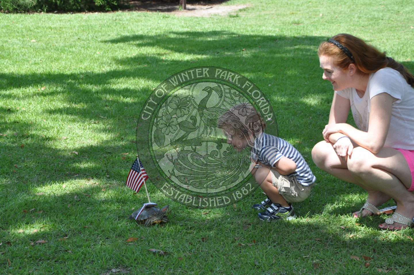An female adult with a young boy squatting down low looking at a turtle with a small American Flag attached to this shell