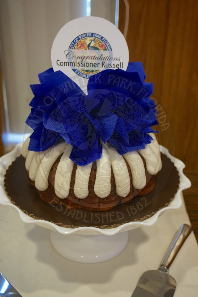 Carrot bundt cake with buttercream frosting. There is a blue paper flower on top of the bundt cake with a paper in the middle . On the paper is the City of Winter Park city seal that says congratulations, Commissioner Russell.