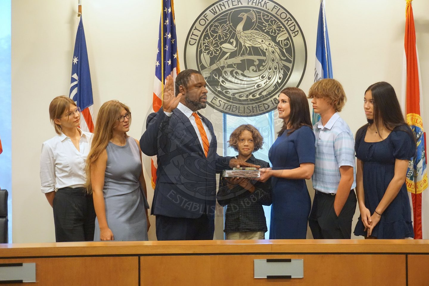 Commissioner Russell holding his left hand on the bible and right hand up in the air. His wife is holding the bible in front of him with the 5 kids surrounding them on the dias.
