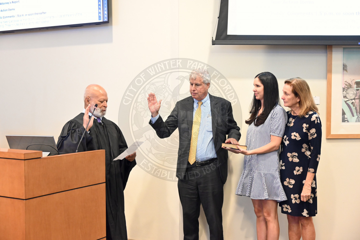 a judge holding his right hand up on a paper while commissioner has his left hand on a bible and his right hand in the air with two females standing next to him.