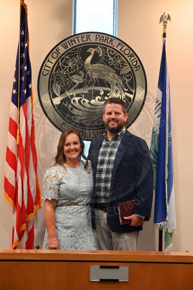 A smiling man and woman stand in front of the City of Winter Park seal and two flags inside an official building.