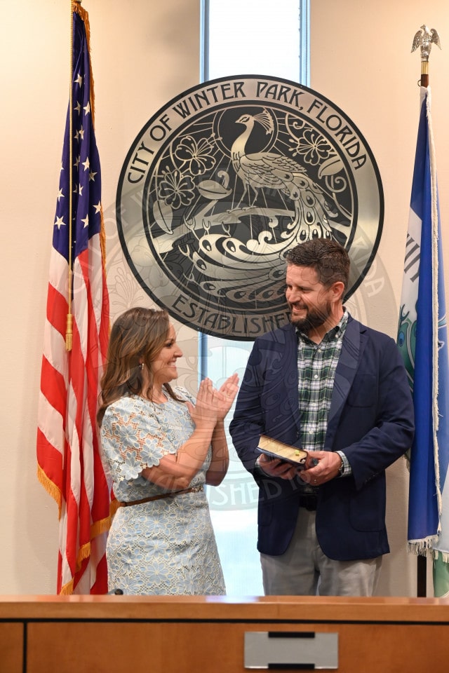 A woman claps as a man holding a book smiles as they stand in front of flags and a City of Winter Park seal..