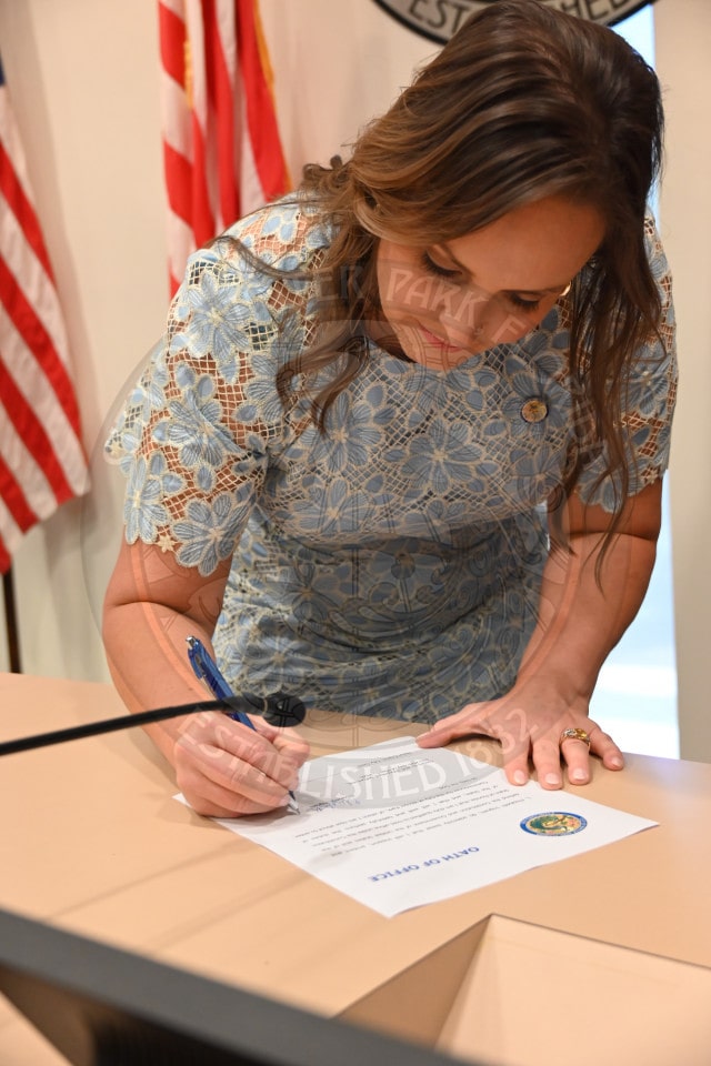 A woman in a blue lace dress signs an official document at a desk with USA flags in the background