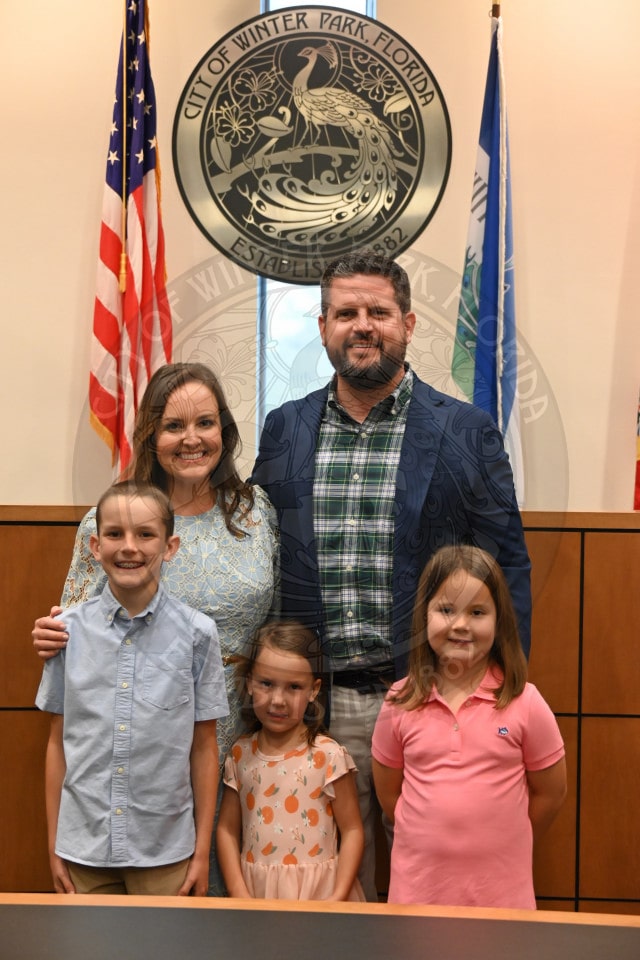 A family of five poses and smiles in front of a City of Winter Park seal and two flags.