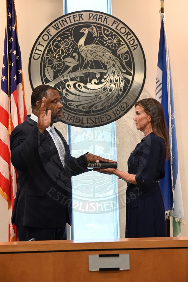 A man is sworn in with his right hand raised, standing beside a woman holding a Bible, in front of Winter Park, Florida seal.