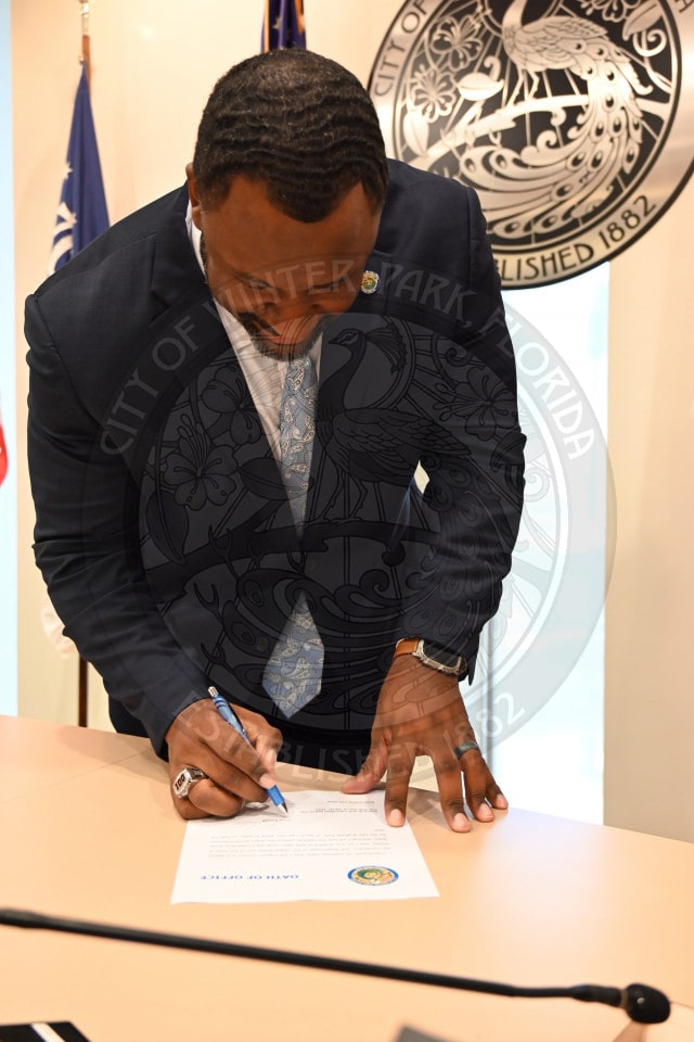A man in a suit signs an official document at a desk with USA flags in the background