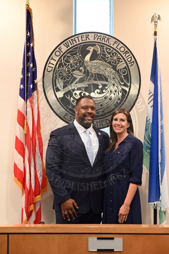 A man and woman stand smiling in front of a City of Winter Park seal between two flags in an official setting.