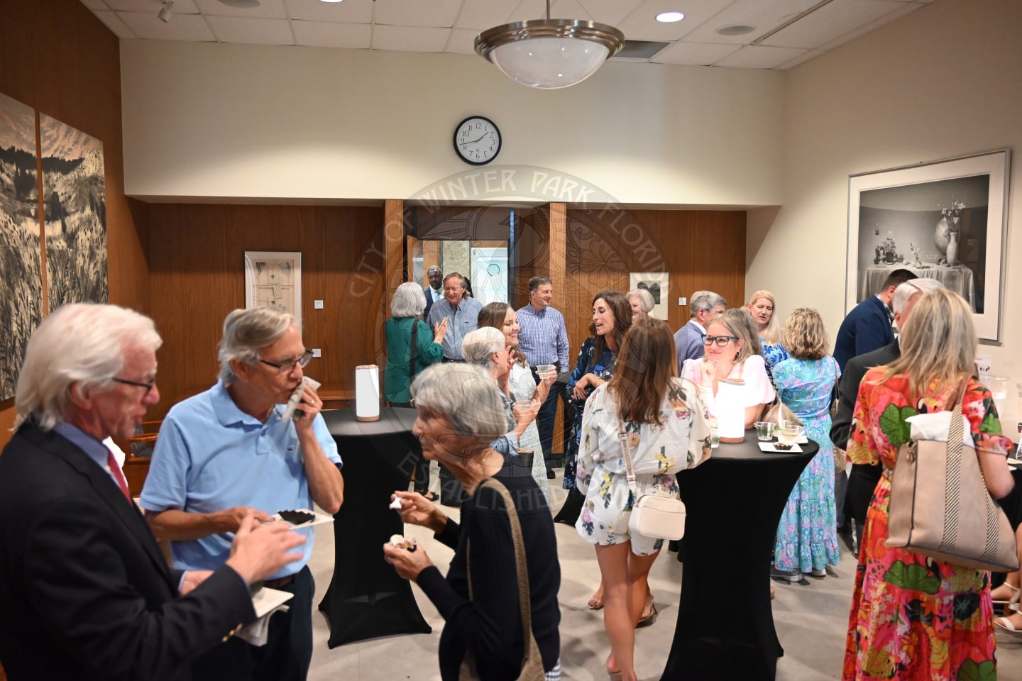 People mingle and chat at a social event in a room with art on the walls and a clock above the door.