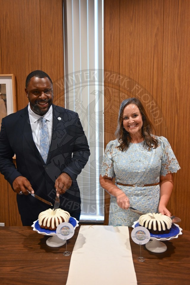A man and woman smile while standing behind two bundt cakes, preparing to cut them on a table.