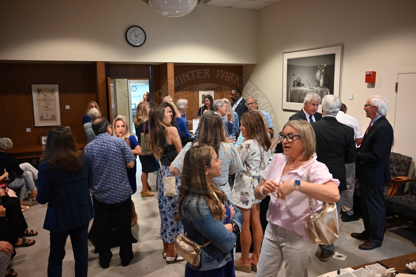People mingle and chat at a social event in a room with art on the walls and a clock above the door.