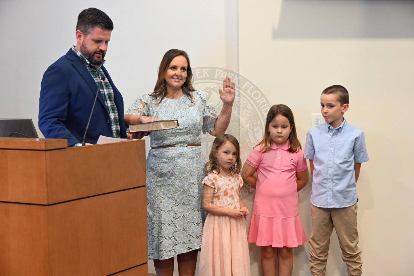 A woman takes an oath with her hand on a Bible, standing beside a man and three children at a podium.