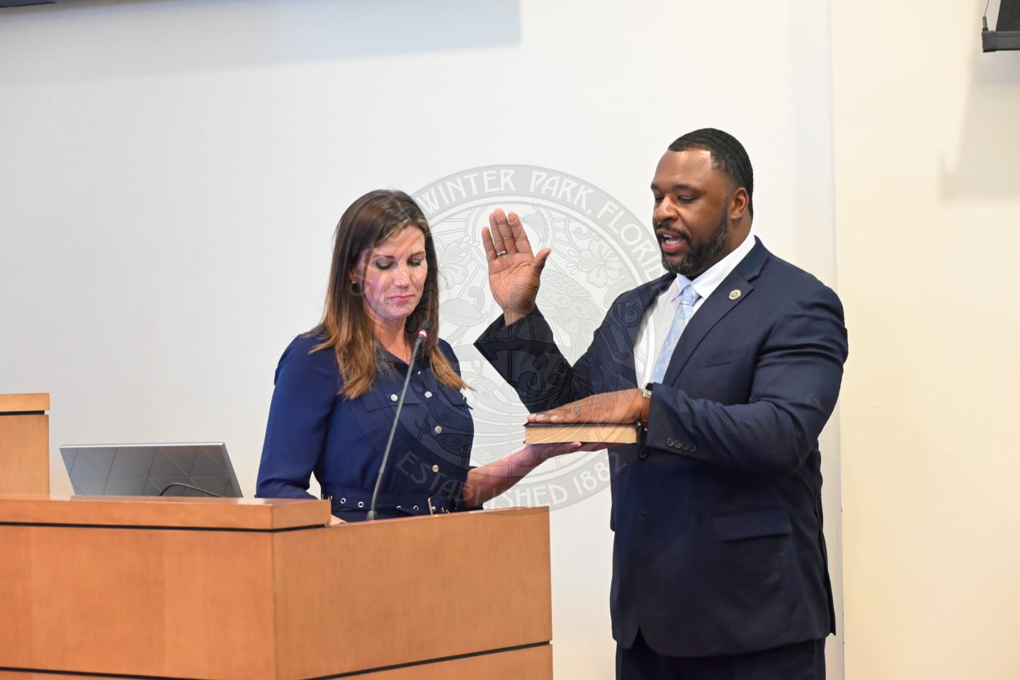 A man in a suit is sworn in with his hand raised, while a woman holds a Bible for him at a podium.