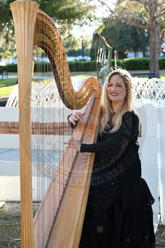 A smiling woman in a black dress plays a large harp outdoors near a fountain and greenery on a sunny day.