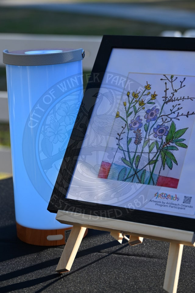A framed floral artwork on an easel is displayed next to a glowing blue cylindrical lamp on a table, with a seal reading City of Winter Park visible in the background.