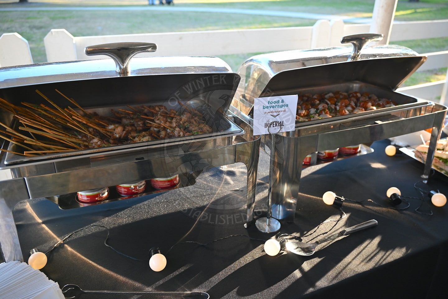 Two metal buffet trays on a black tablecloth, one filled with skewered meat and the other with small appetizers. A small sign reads Food &amp; Beverages. String lights are draped across the table.