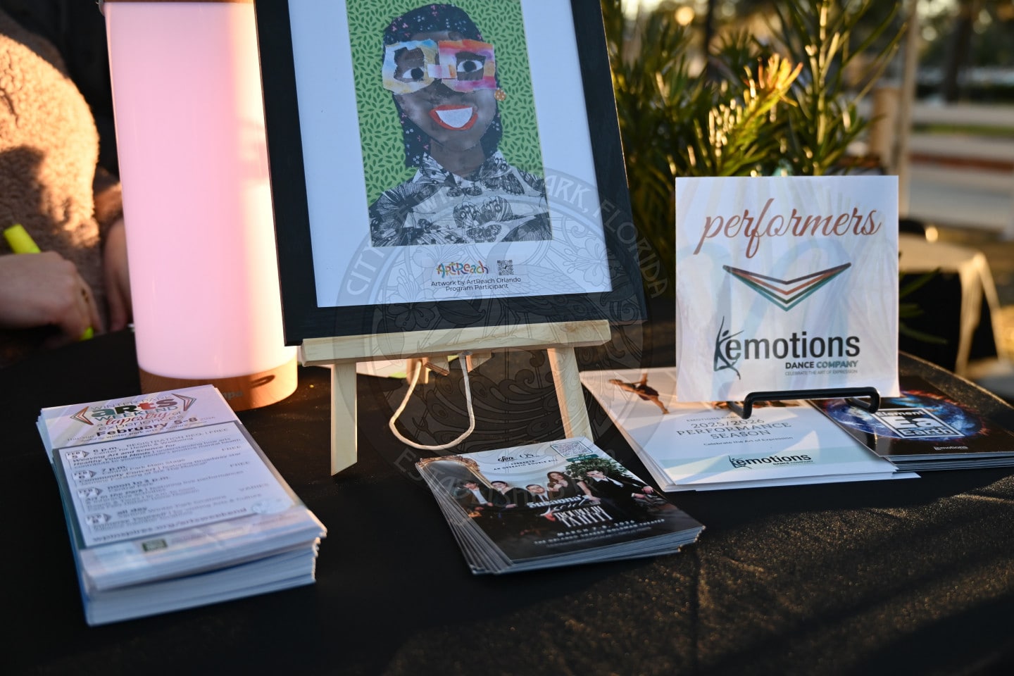 A table displays art show flyers, a booklet, a program for Emotions Dance Company, and a colorful portrait on an easel. Soft lighting and plants are visible in the background.