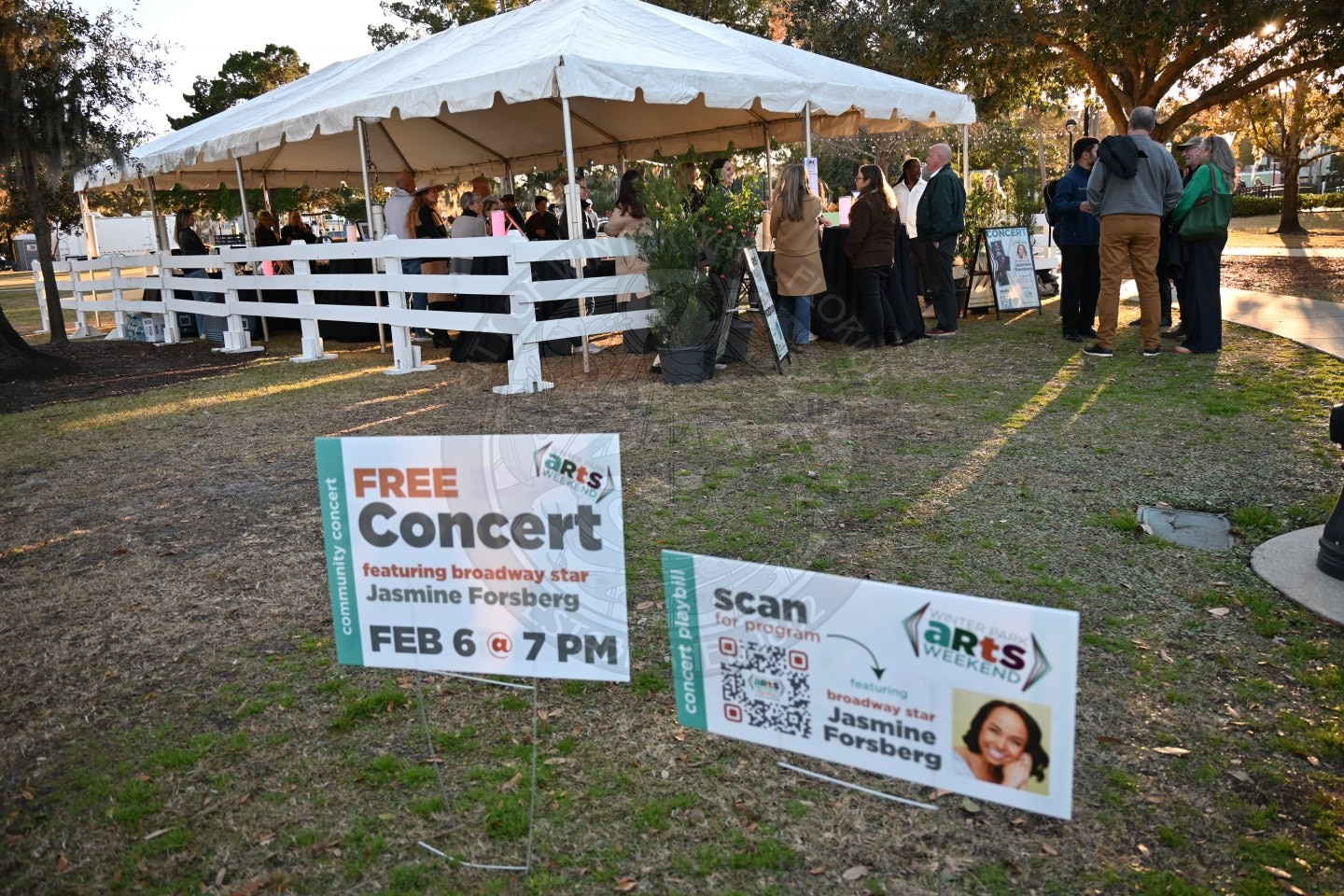 People gather near a white tent in a park for a free concert event. In the foreground, yard signs advertise the concert featuring Broadway star Jasmine Forsberg on February 6 at 7 PM, with QR codes for more information.