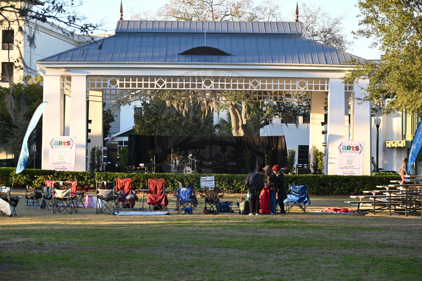 Outdoor stage with a metal roof, empty chairs arranged in front, a few people standing nearby, and banners reading ARTS Weekend on each side. The setting appears to be a park with trees and open grassy area.