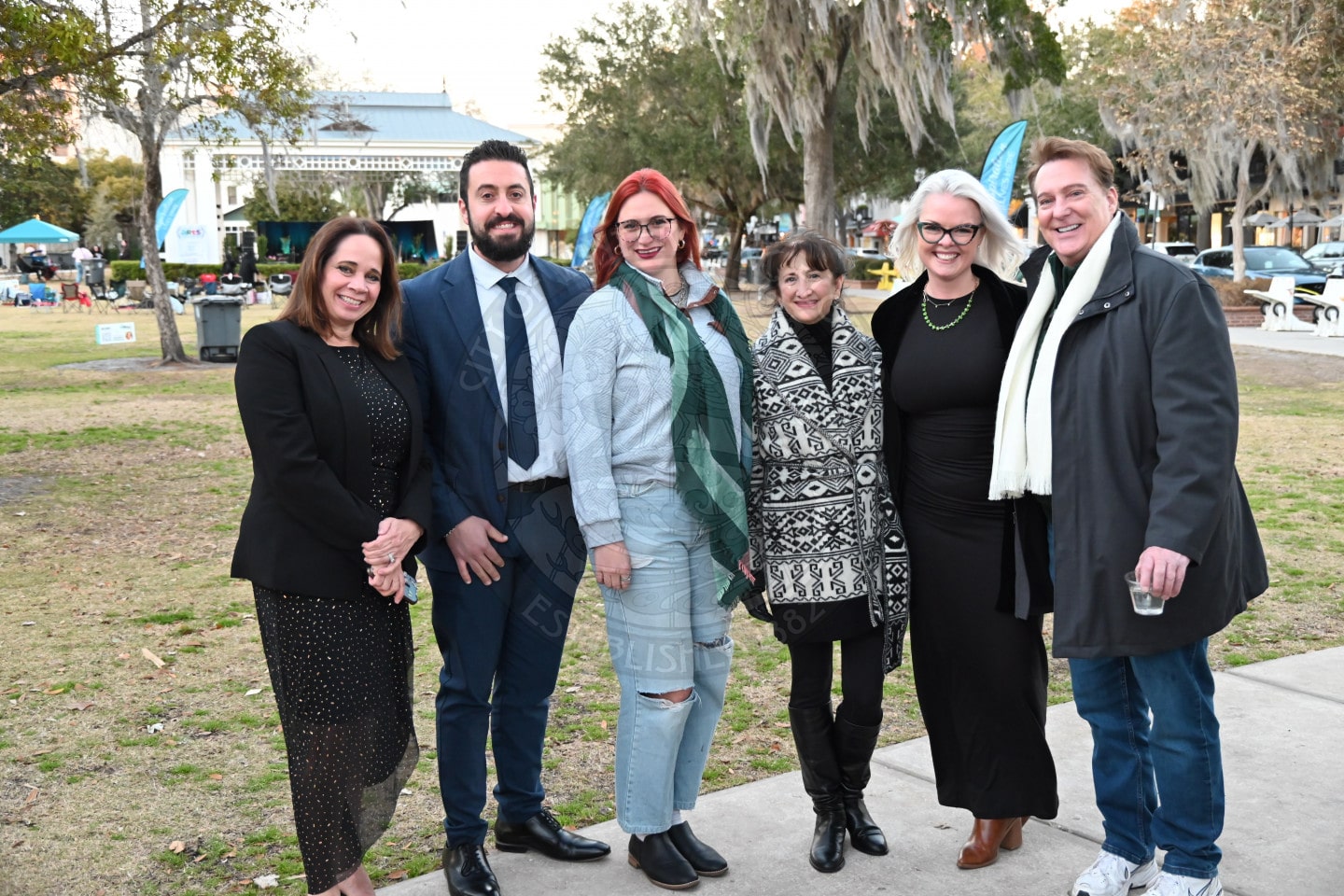 Six adults pose and smile together outdoors in a park. They are dressed in winter clothing, including coats and scarves. Trees and tents are visible in the background.