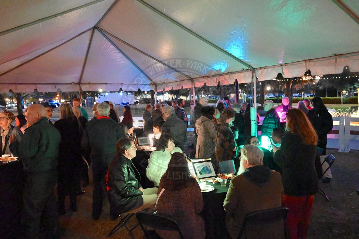 A large group of people gather under a white event tent at night, socializing and looking at tables with displays. Colorful lights illuminate the area, creating a festive atmosphere.